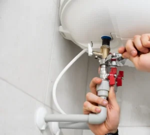 Close-up van iemands handen bij het installeren of repareren van leidingen en kleppen onder een witte boiler, onderdeel van een Hybride Warmtepomp installatie, gemonteerd op een betegelde muur.