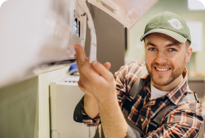 Een glimlachende technicus met een groene pet en een geruit overhemd werkt aan een airconditioner en past met zijn handen onderdelen aan. Dit is een demonstratie van de expertise van Yas Installatie.