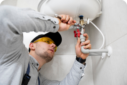 Een loodgieter van Yas Installatie met een veiligheidsbril op en een pet op gebruikt een moersleutel om leidingen te repareren die zijn aangesloten op een boiler die aan een muur is bevestigd.