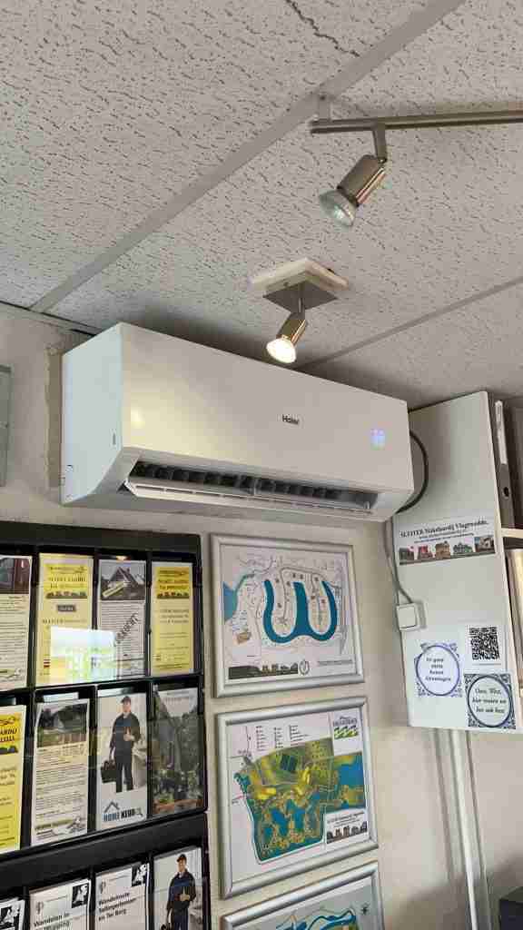 Een witte Haier airconditioner aan de muur is geïnstalleerd op een plafond met structuur boven ingelijste posters, kaarten en informatiebrochures in een goed verlichte kamer. In de buurt van de airconditioner is een spot gemonteerd.