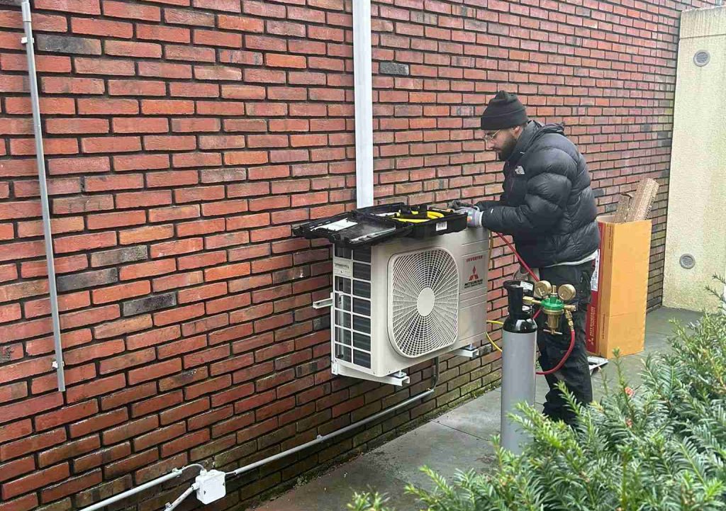 Een persoon in een zwarte jas en muts werkt aan een buitenairco boven Yas Installatie, gemonteerd op een bakstenen muur, met gereedschap en een gasfles in de buurt.