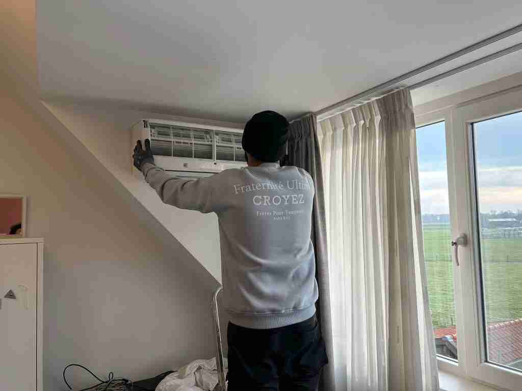 Een persoon in een grijs sweatshirt en zwarte muts voert een airco-installatie uit op een unit boven een raam in een lichte kamer met witte muren en grote gordijnen.