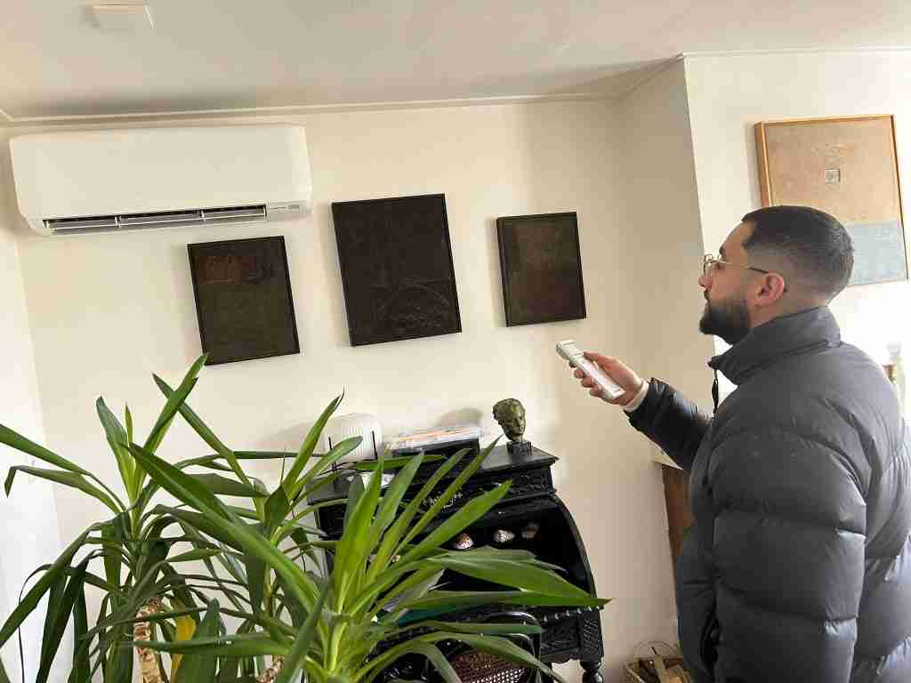 Een man met een bril en een gewatteerd jack gebruikt een afstandsbediening om een airco-installatie te bedienen die aan de muur is bevestigd in een woonkamer met planten, kunstwerken en planken.