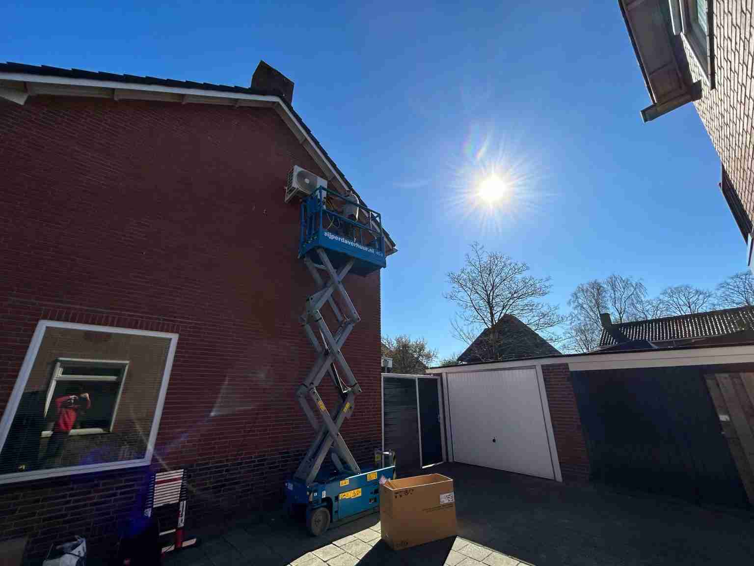 Een persoon op een blauwe schaarhoogwerker voert een hybride warmtepompinstallatie uit of onderhoudt een airconditioningunit aan de zijkant van een bakstenen huis op een zonnige dag, met een heldere hemel en een kartonnen doos op de grond vlakbij.