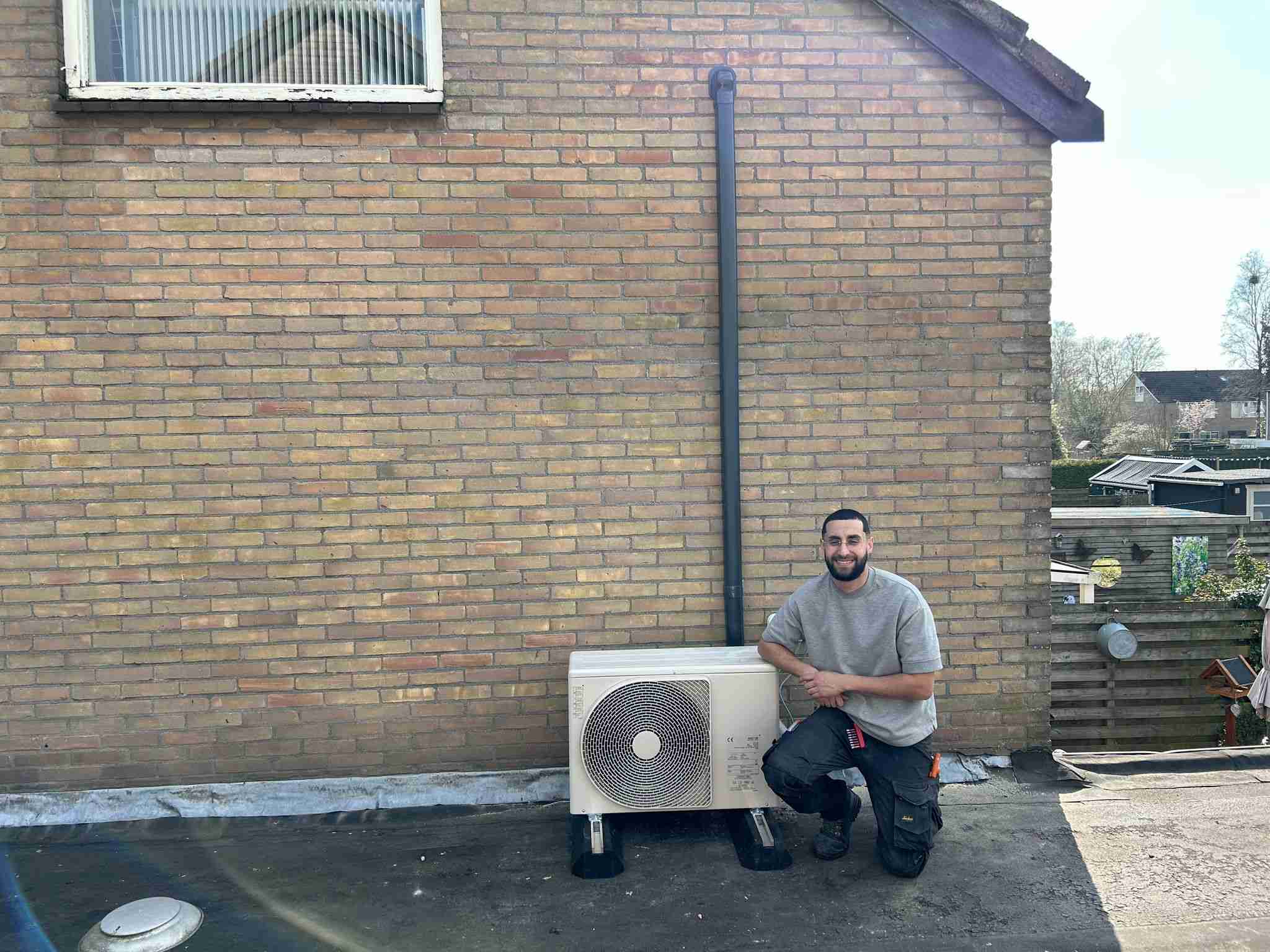 Een man knielt op een dak naast een gemonteerde hybride warmtepompinstallatie die aan een bakstenen muur is bevestigd en lacht naar de camera. Het is een zonnige en heldere dag.