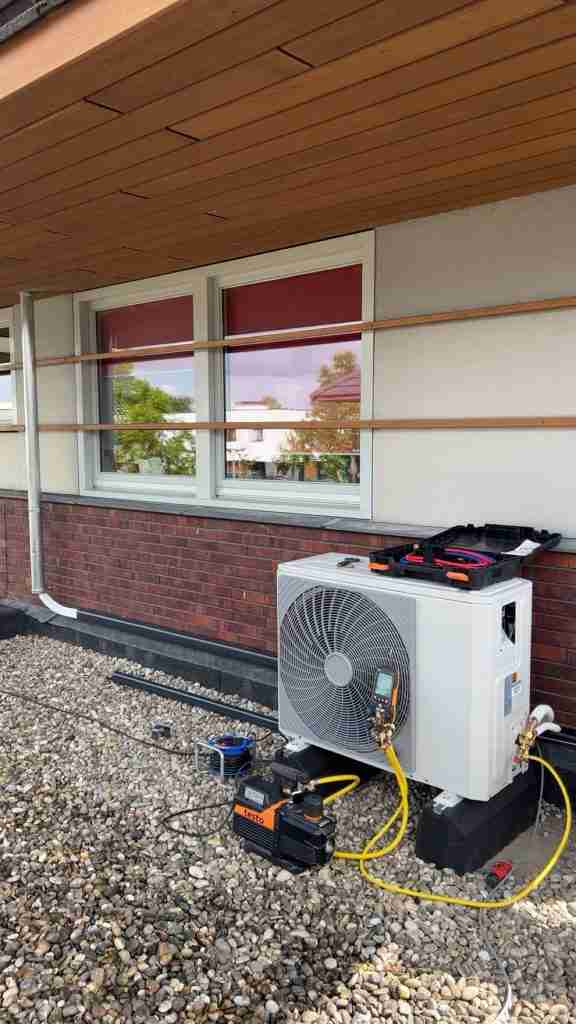 Een airco installatie-eenheid staat opgesteld buiten een gebouw met bakstenen en bakstenen muren. Gele kabels en gereedschap zijn bevestigd aan de unit, die op grind rust onder twee ramen met rode jaloezieën.