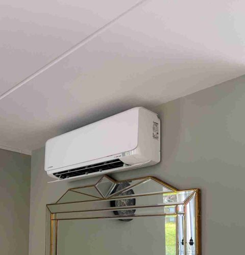 Een witte wandairco is geïnstalleerd boven een decoratieve spiegel op een lichtgrijze muur in een kamer met een wit plafond.