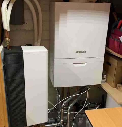 Een modern boilersysteem met geïsoleerde leidingen, een witte wandgemonteerde ATAG-unit en een hybride warmtepompinstallatie, allemaal netjes gerangschikt in een nette bijkeuken met planken en opbergdozen.