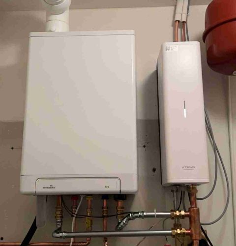 Een witte wandketel met aangesloten leidingen en kleppen maakt deel uit van een hybride warmtepompinstallatie, geïnstalleerd naast een hoog, slank wit elektronisch apparaat en een grote rode tank in een bijkeuken.