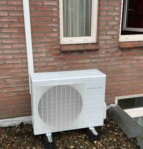 Een witte hybride warmtepomp installatie-unit is geïnstalleerd buiten een bakstenen gebouw onder een raam, rustend op zwarte steunen op een grindoppervlak met een witte afvoerpijp die er verticaal naast loopt.