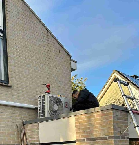 Een persoon in een donkere jas werkt aan een buitenairco boven Yas Installatie, vastgemaakt aan een bakstenen gebouw, met een ladder tegen de muur en huizen zichtbaar op de achtergrond onder een gedeeltelijk bewolkte hemel.