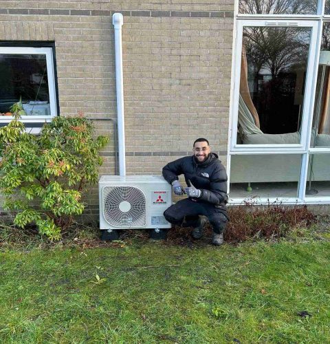 Een persoon in donkere kleding knielt en glimlacht naast een buitenairco die op een bakstenen muur is gemonteerd en trots zijn werk laat zien over Yas Installatie, met gras, een struik en een groot raam in de buurt.