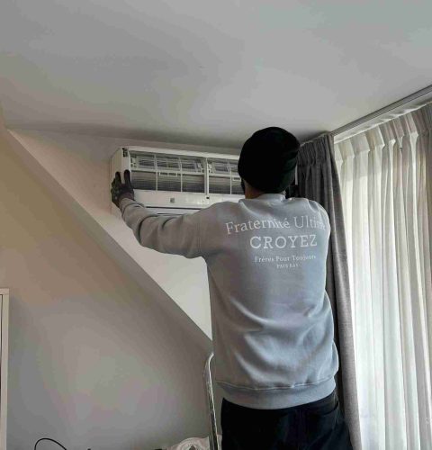 Een persoon met een muts en een sweatshirt voert een airco-installatie uit, waarbij hij een airconditioningsunit aan de muur onderhoudt in een lichte kamer met gordijnen en een groot raam.
