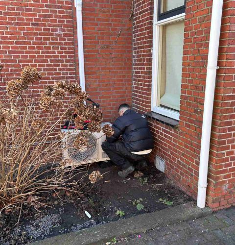 Een persoon in een zwarte jas hurkt naast een bakstenen gebouw, terwijl hij een airco-installatie uitvoert op een buitenunit in de buurt van gedroogde hortensiastruiken.