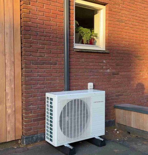 Een witte hybride warmtepompinstallatie staat tegen een rode bakstenen muur onder een raam met een plant erin. Grijze leidingen lopen verticaal van de unit tot boven het raam en de grond lijkt bestraat.