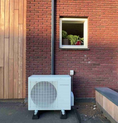 Een hybride warmtepompinstallatie staat tegen een rode bakstenen muur onder een klein raam met potplanten. Een zwarte pijp loopt verticaal vanaf de unit en buigt er horizontaal bovenuit.