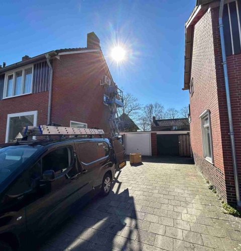 Een zwarte bestelwagen met ladders erop staat geparkeerd naast een bakstenen huis terwijl een arbeider op een lift een hybride warmtepompinstallatie installeert bij het dak onder helder zonlicht en een strakblauwe lucht.