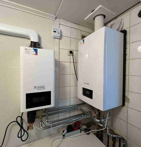 Twee witte wandgemonteerde verwarmingsunits met digitale displays, onderdeel van een hybride warmtepompinstallatie, zijn geïnstalleerd in een betegelde bijkeuken, verbonden door buizen en draden, met een ontluchtingspijp naar buiten.