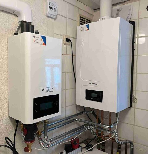 Twee witte wandverwarmingsunits, die deel uitmaken van een hybride warmtepompinstallatie, hebben digitale displays en zijn verbonden door verschillende leidingen in een betegelde bijkeuken. Erboven is een klein bedieningstoestel gemonteerd.